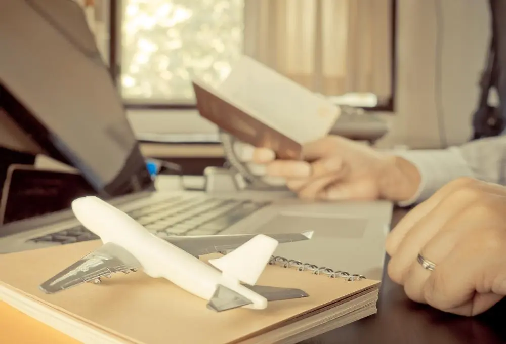 Человек сидит за столом с паспортом в руках; на столе стоит ноутбук и модель пассажирского самолета